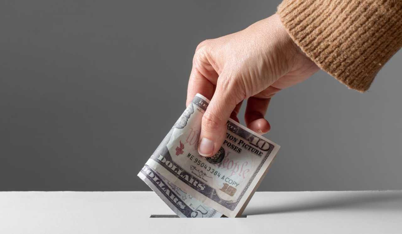 A stock photo of a hand placing money into a donation box. A stock photo of a hand placing money into a donation box.