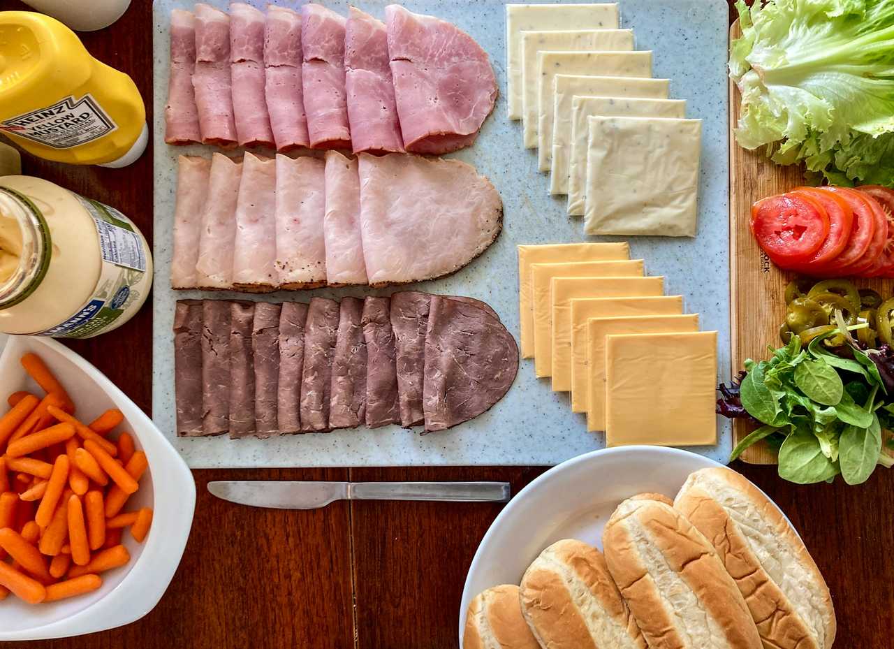 A variety of sandwich meats, cheese, bread, vegetables, and condiments on a table. A variety of sandwich meats, cheese, bread, vegetables, and condiments on a table.